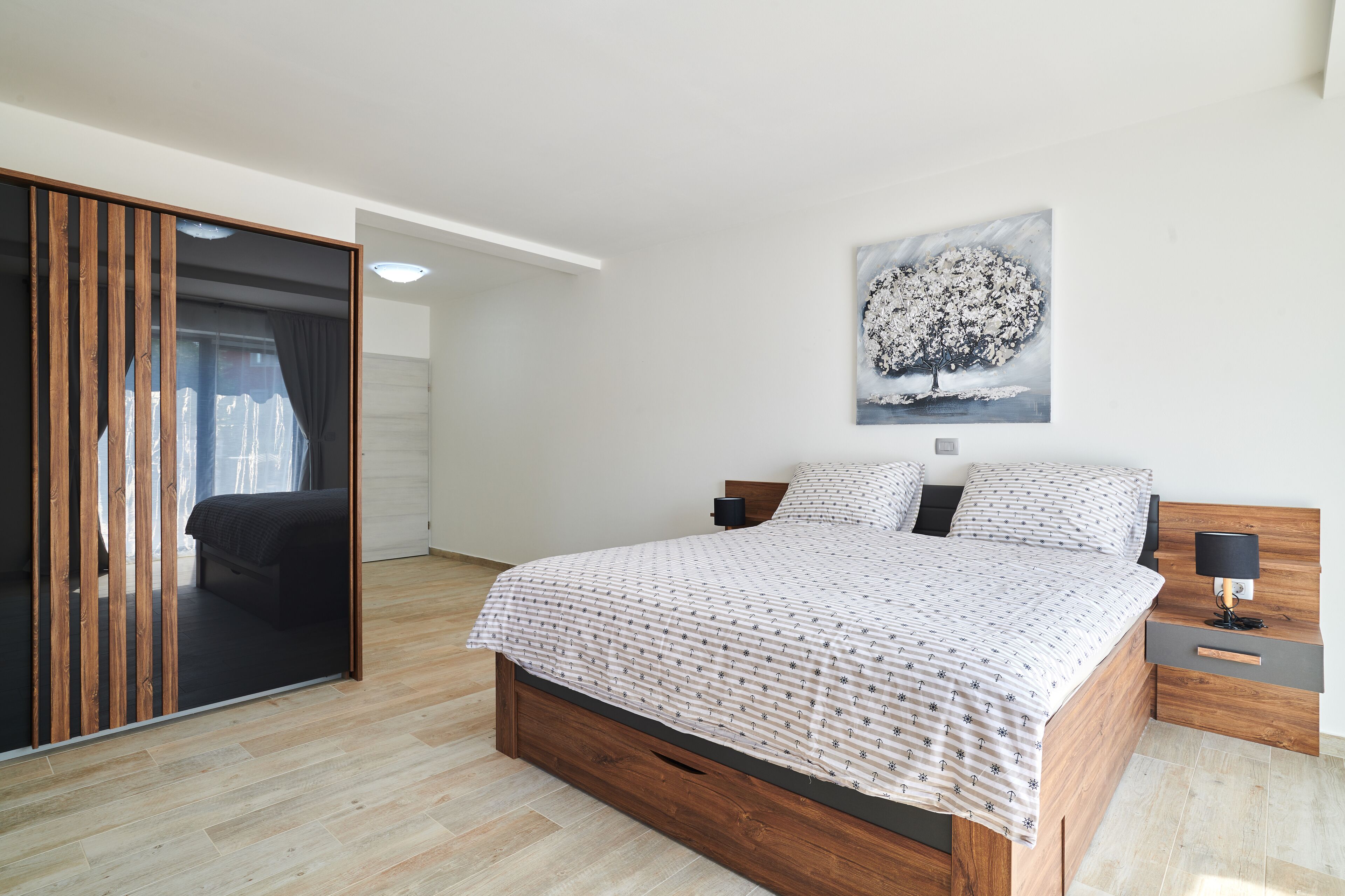 Ein modernes Schlafzimmer mit einem hölzernen Bettgestell, einem großen Fenster mit Vorhängen, einem Baumgemälde und einem Nachttisch mit einer Lampe.