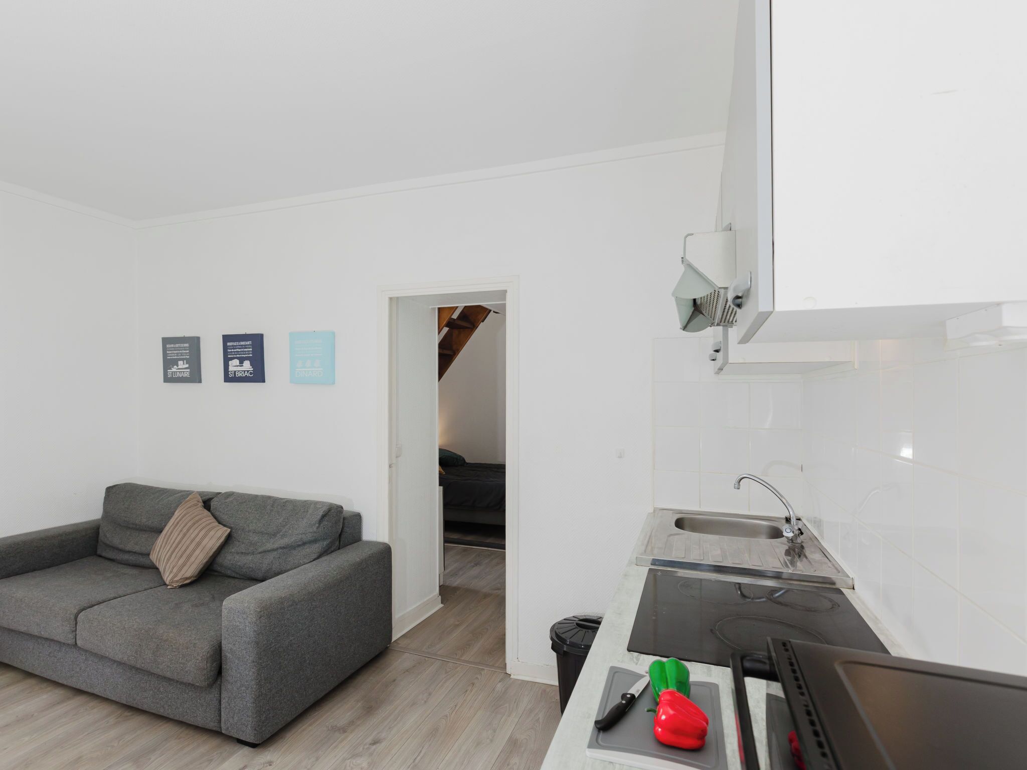 A modern apartment with a grey sofa, a kitchen with a sink and stove, and a hallway leading to a staircase.