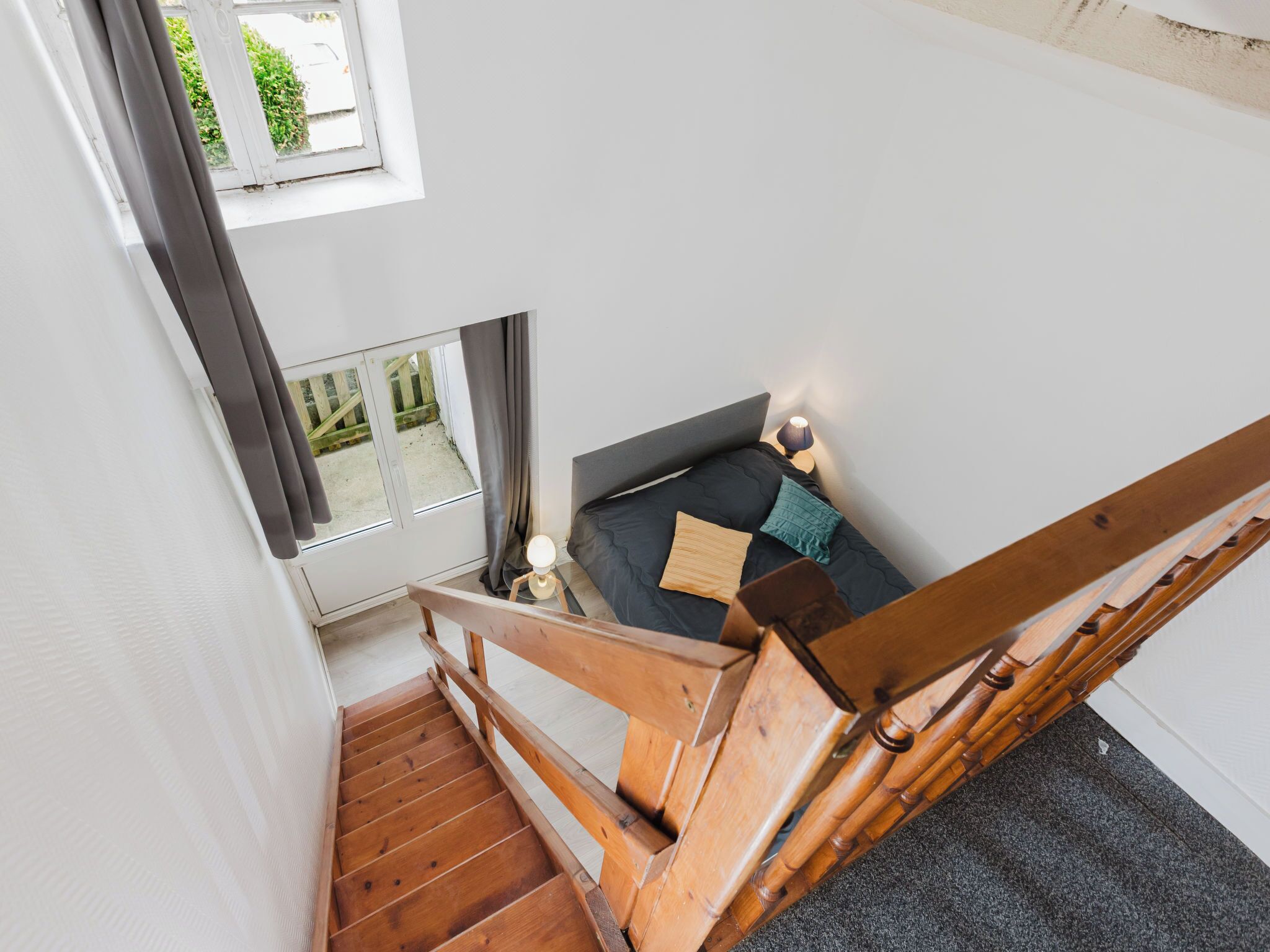 Ein Schlafzimmer mit Bett, Nachttisch, Lampe, einem Fenster mit Vorhängen und einer Holztreppe.