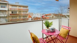 Uma varanda com toldo listrado, uma mesa pequena com cadeiras, uma planta em vaso e vista de edifícios ao fundo.