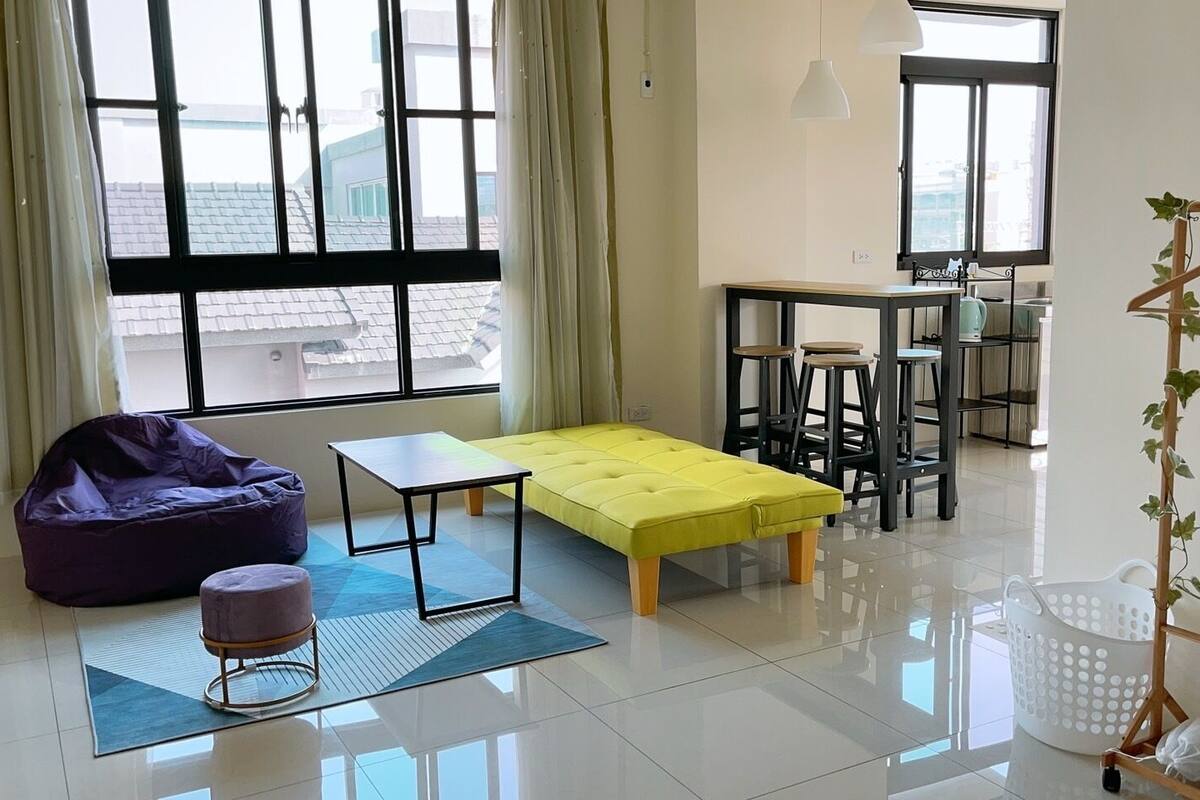 Un salon moderne avec un fauteuil poire violet, un pouf jaune, une petite table et une plante suspendue.