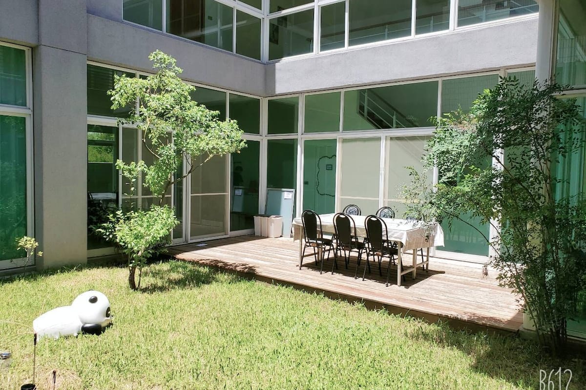 Un bâtiment moderne doté de grandes fenêtres, d’un patio avec une table et des chaises, et d’un petit arbre.