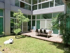 Un edificio moderno con amplios ventanales, un patio con mesa y sillas, y un pequeño árbol.