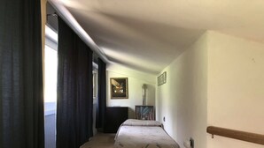 Une chambre d’hôtel étroite, avec un lit, une table de chevet, un tableau au mur et une fenêtre avec des rideaux.