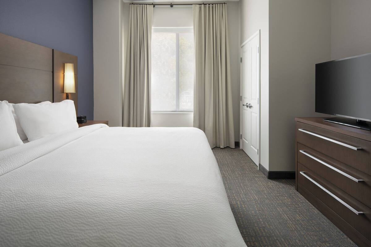 A hotel room with a large bed, a television, a nightstand, and a window with curtains.