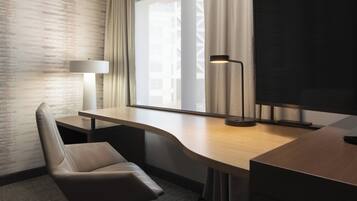 Quarto de hotel moderno com escrivaninha curva, poltrona confortável, janela grande com cortinas e TV de tela plana.