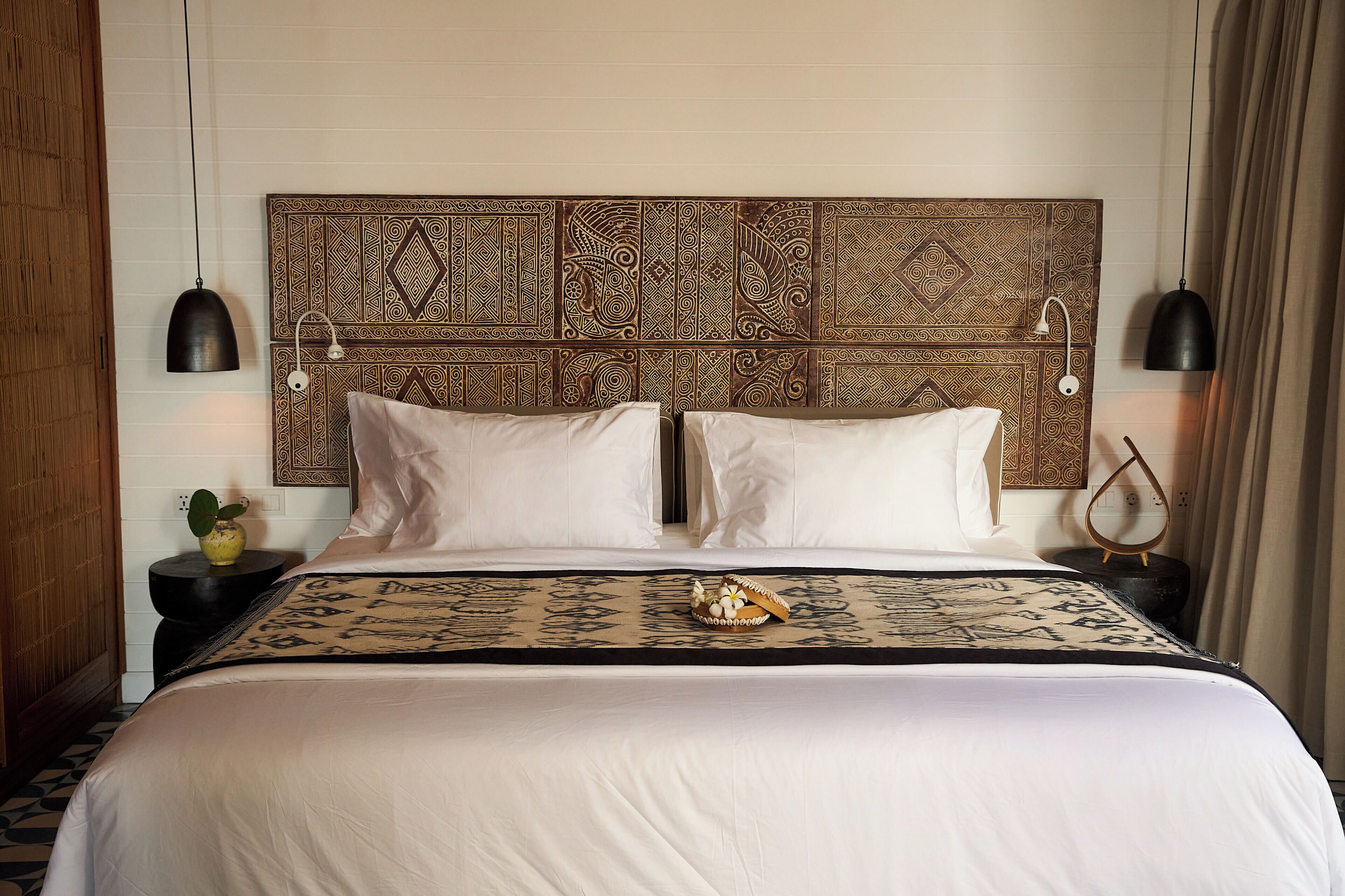 Una cama bien hecha con un cabecero estampado, dos mesitas de noche con lámparas y un jarrón decorativo sobre una de ellas.