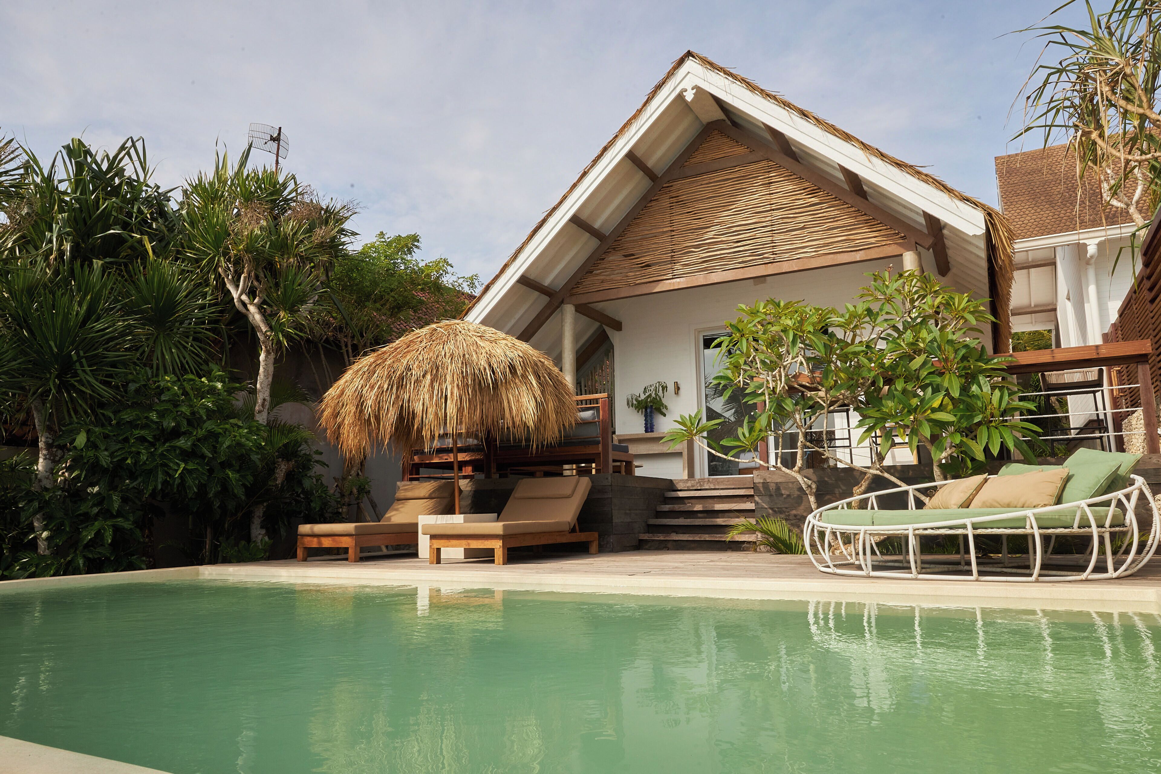 A poolside area with a thatched umbrella, wooden lounge chairs, and a white wicker sofa.