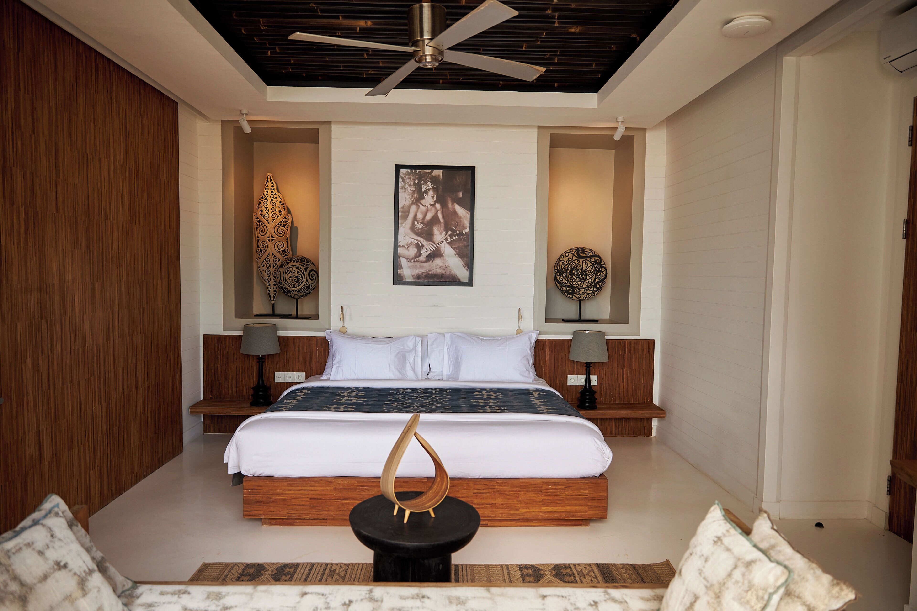 A modern bedroom with a large bed, wooden headboard, two bedside tables with lamps, a ceiling fan, and decorative wall art.
