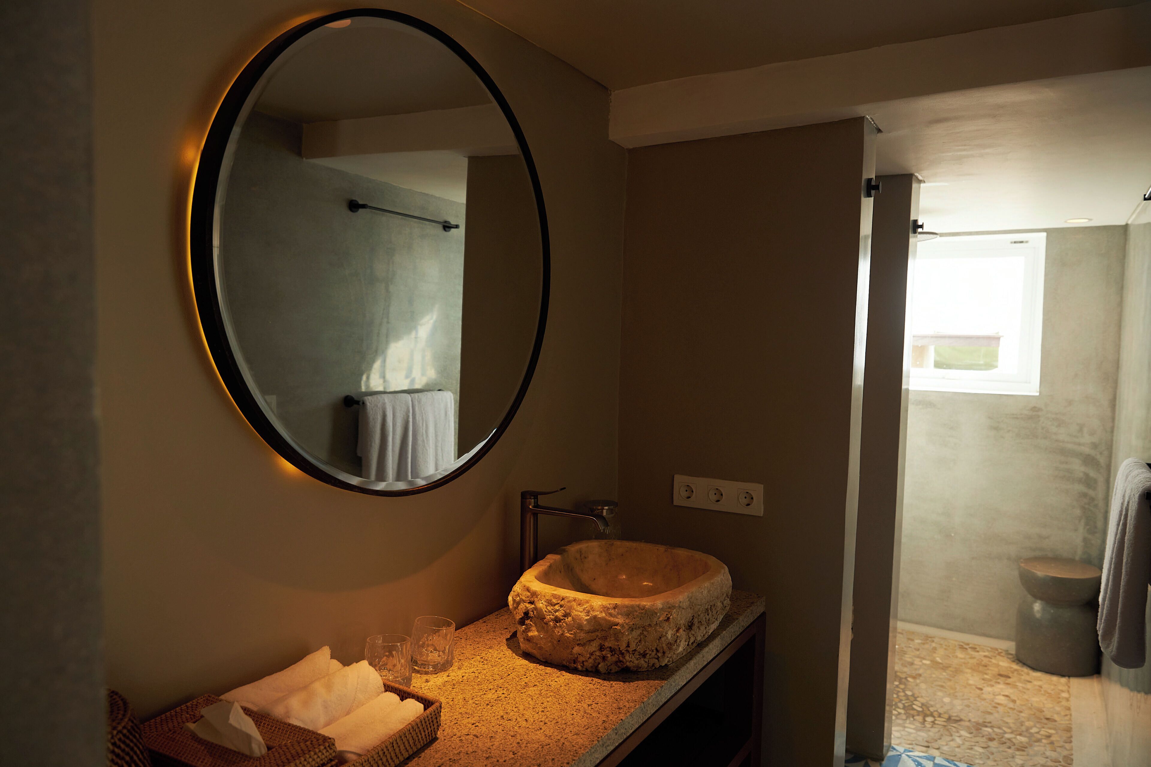 A bathroom with a round mirror, a stone sink, a towel rack, and a shower area.