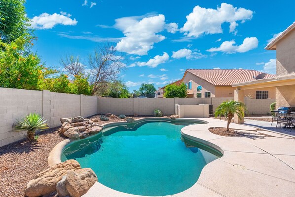 A backyard with a swimming pool, a patio with chairs, and a house with a tiled roof.