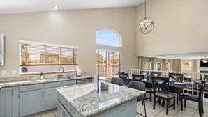 A spacious kitchen with a large island, a dining area with a table and chairs, and a view of the outdoors through large windows. - Brand New Luxury 5BR | Pool, Hot Tub & Mini Golf (Mesa)