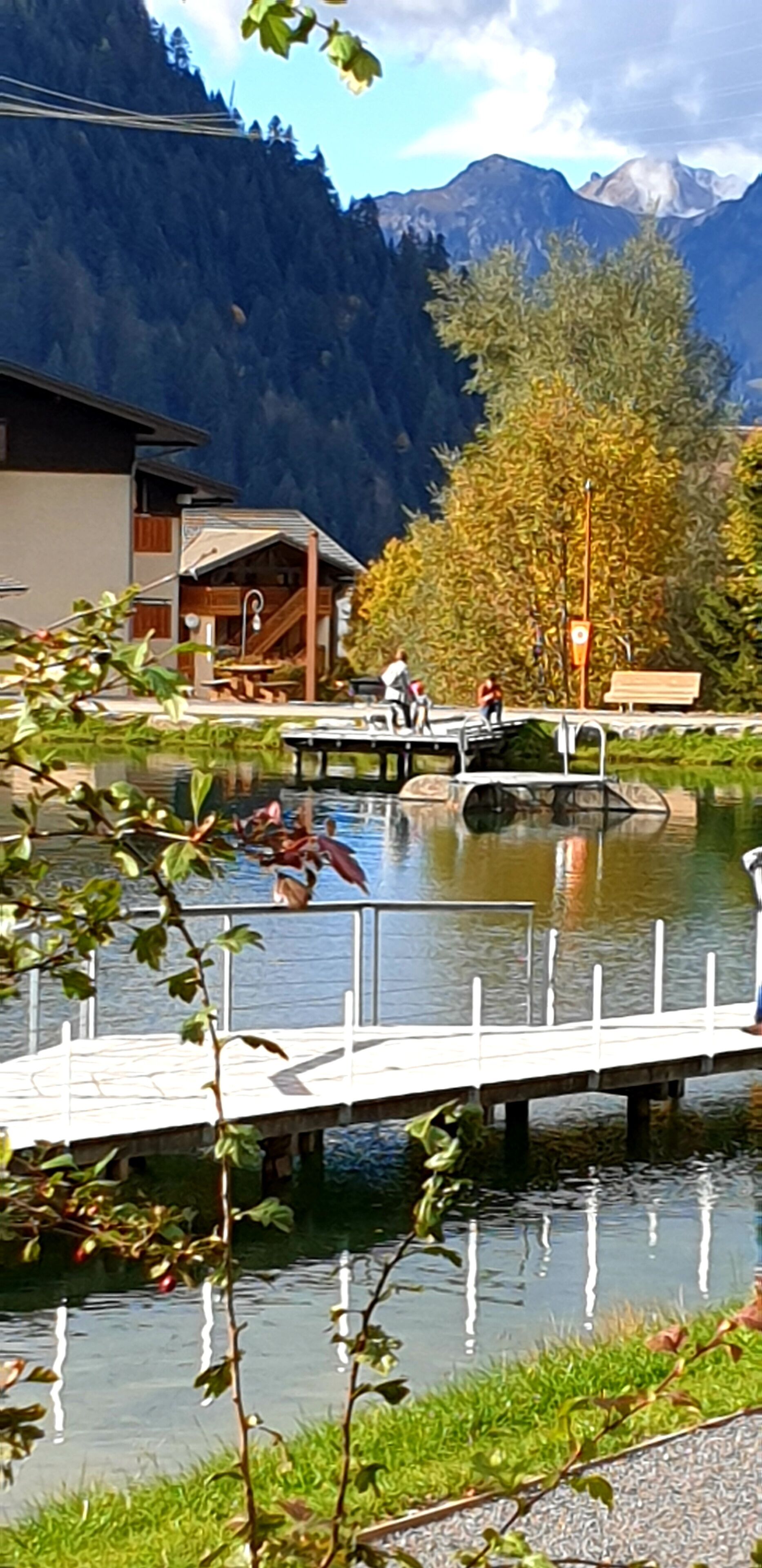 Eine Flusslandschaft mit einer Holzbrücke, einem Gebäude mit schrägem Dach und Bergen im Hintergrund.