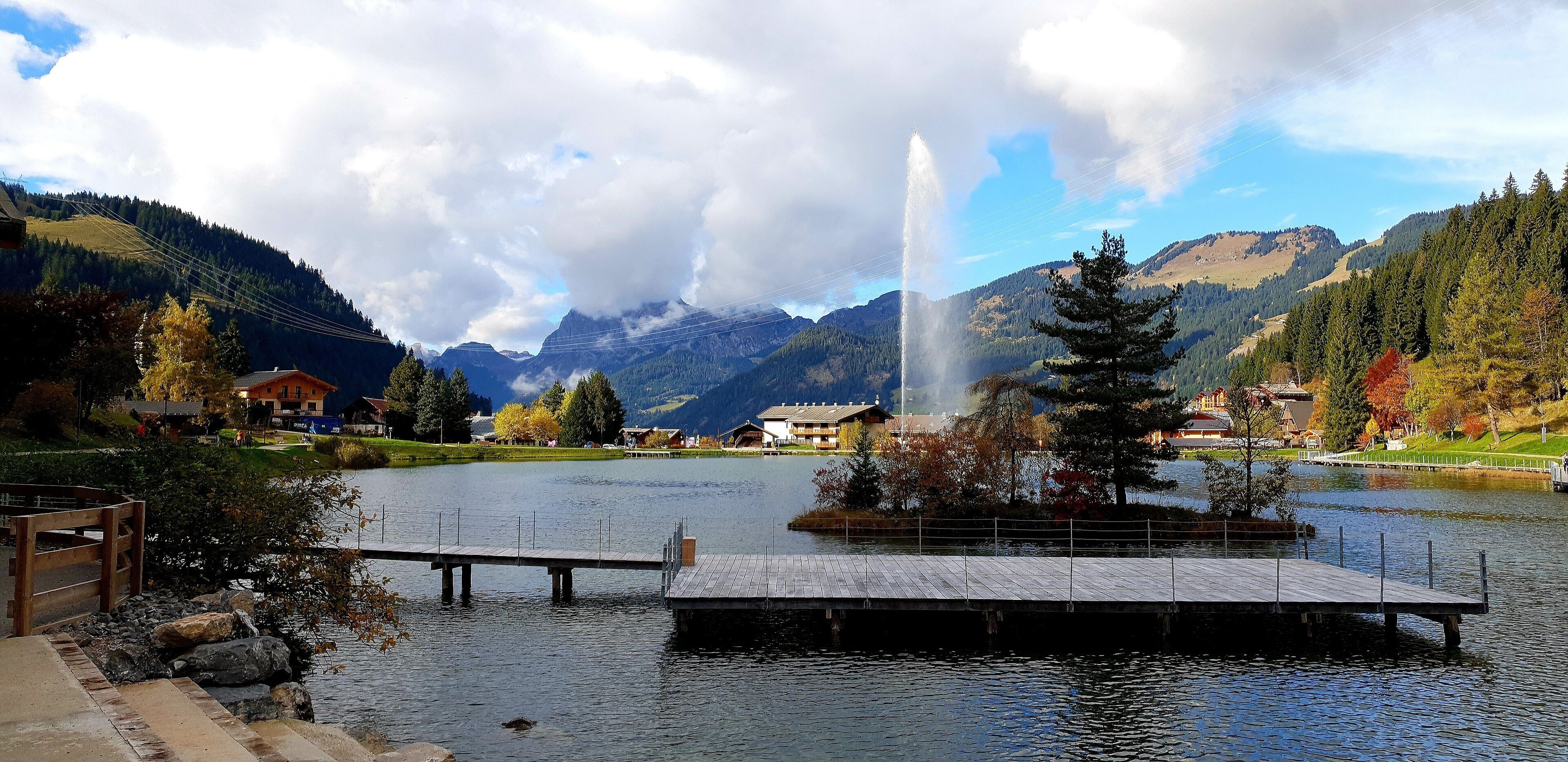 A lake with a wooden dock and a fountain, surrounded by mountains and autumn-colored trees.