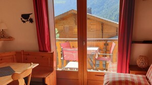 Ein gemütliches Zimmer mit einer hölzernen Sitzbank, einem Tisch und einem Bett mit karierter Bettwäsche. Eine Glastür öffnet sich zu einem Außenbereich mit einem Holztisch und Stühlen.