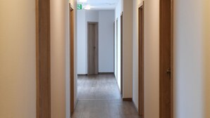 Un pasillo largo y estrecho con puertas de madera a ambos lados, un letrero verde de salida de emergencia y una lámpara en el techo.