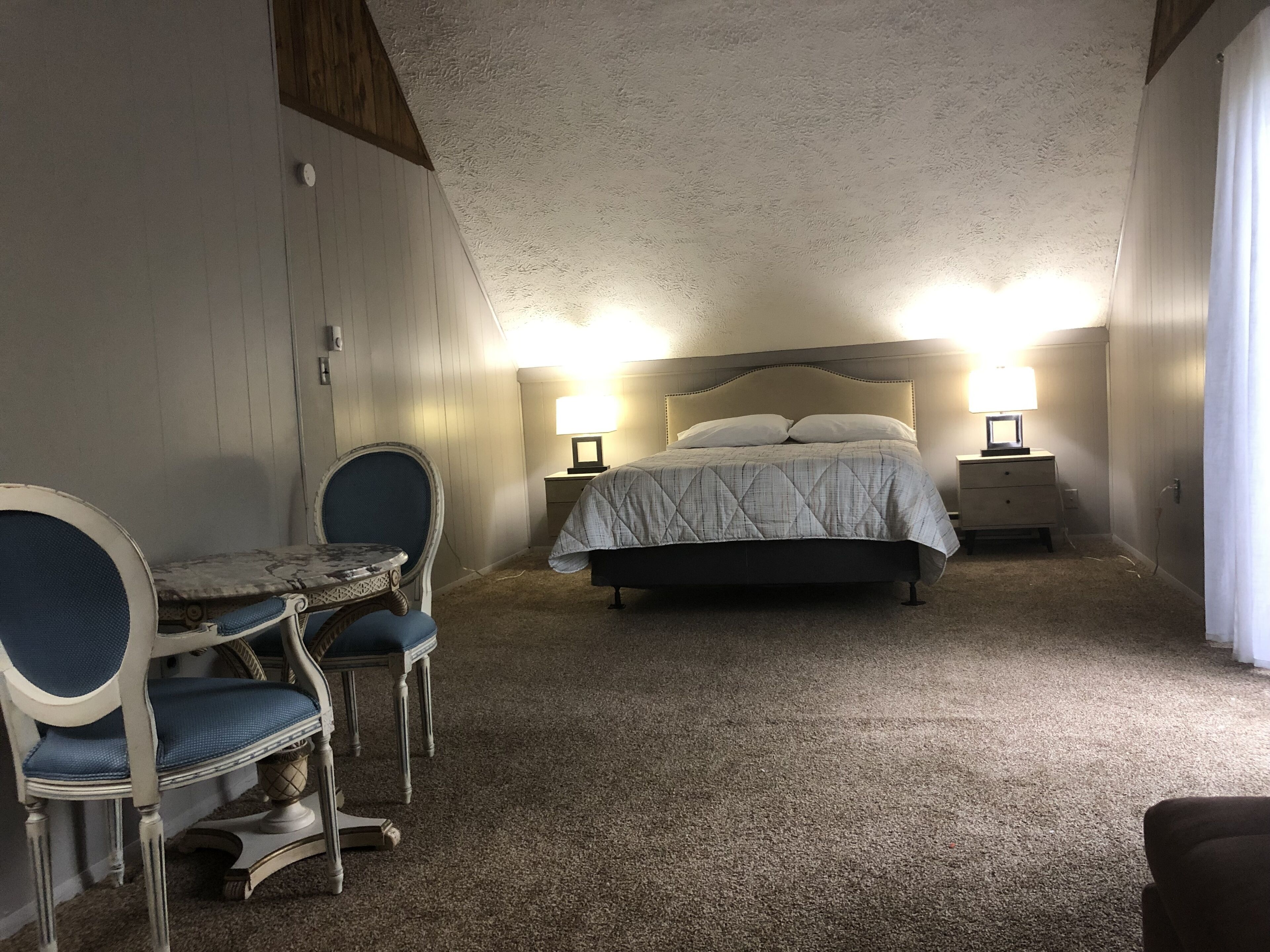 A bedroom with a bed, two bedside tables with lamps, a small table with chairs, and a sloped ceiling.