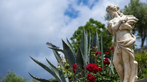 A statue of a figure holding a plant, surrounded by red flowers and large green plants. - Il Mandorlo (Tramonti)