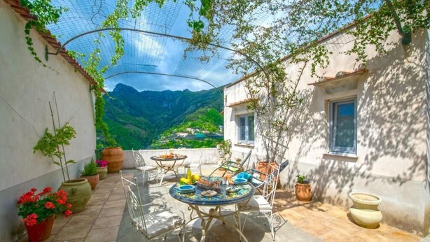 A patio with a round table and chairs, surrounded by potted plants and a view of mountains. - Il Mandorlo (Tramonti)