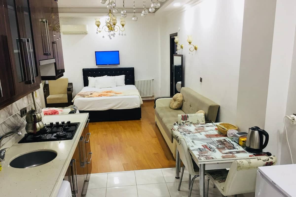 A compact hotel room with a bed, a sofa, a dining table, a kitchen area with a stove, and a television.