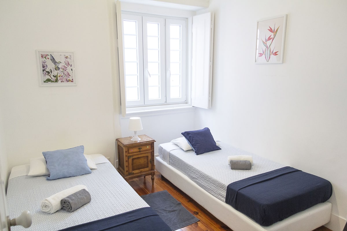 A room with two single beds, a nightstand with a lamp, a window with blinds, and two framed pictures on the wall.