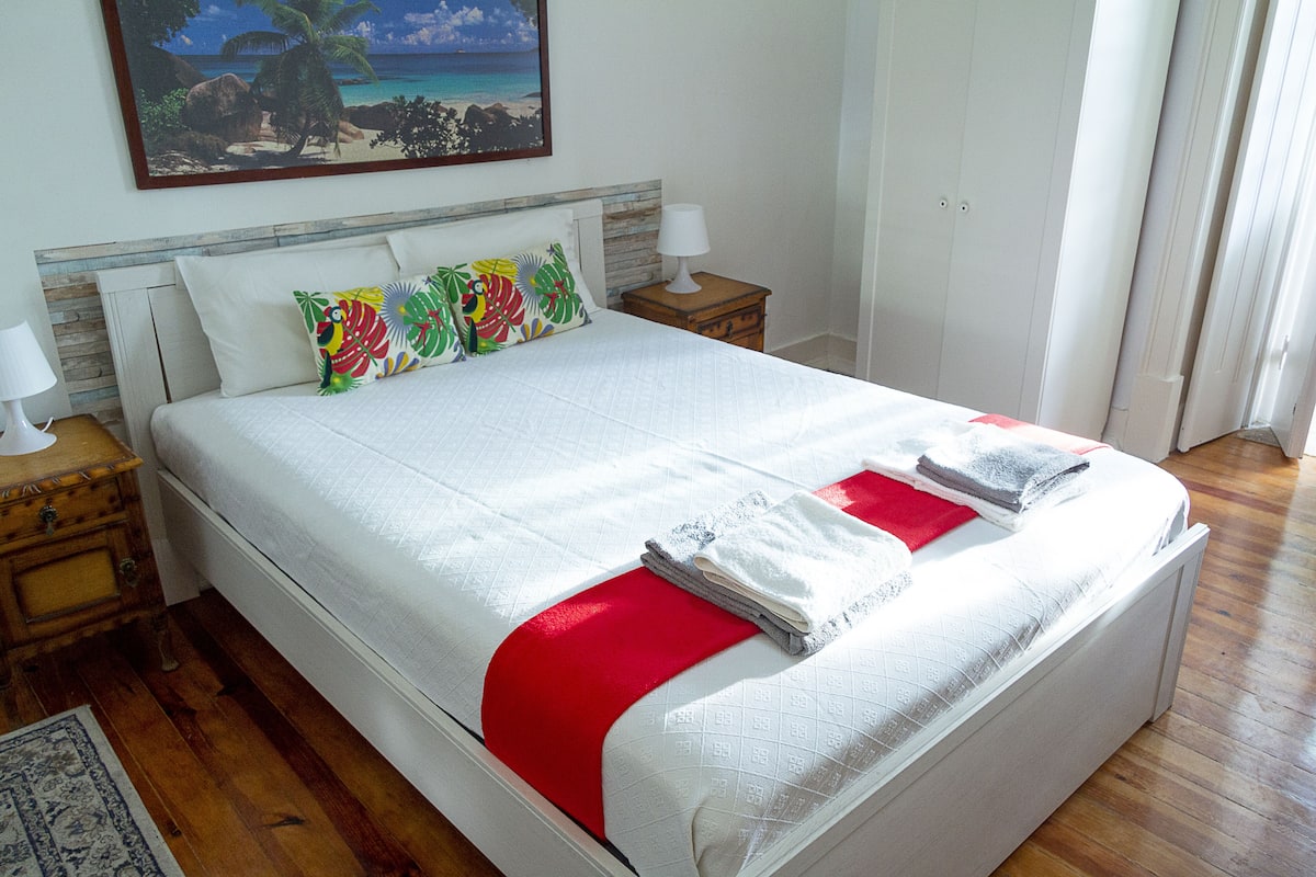 A neatly made bed with white bedding and red accents, a wooden nightstand with a lamp, and a colorful painting of a beach scene on the wall.