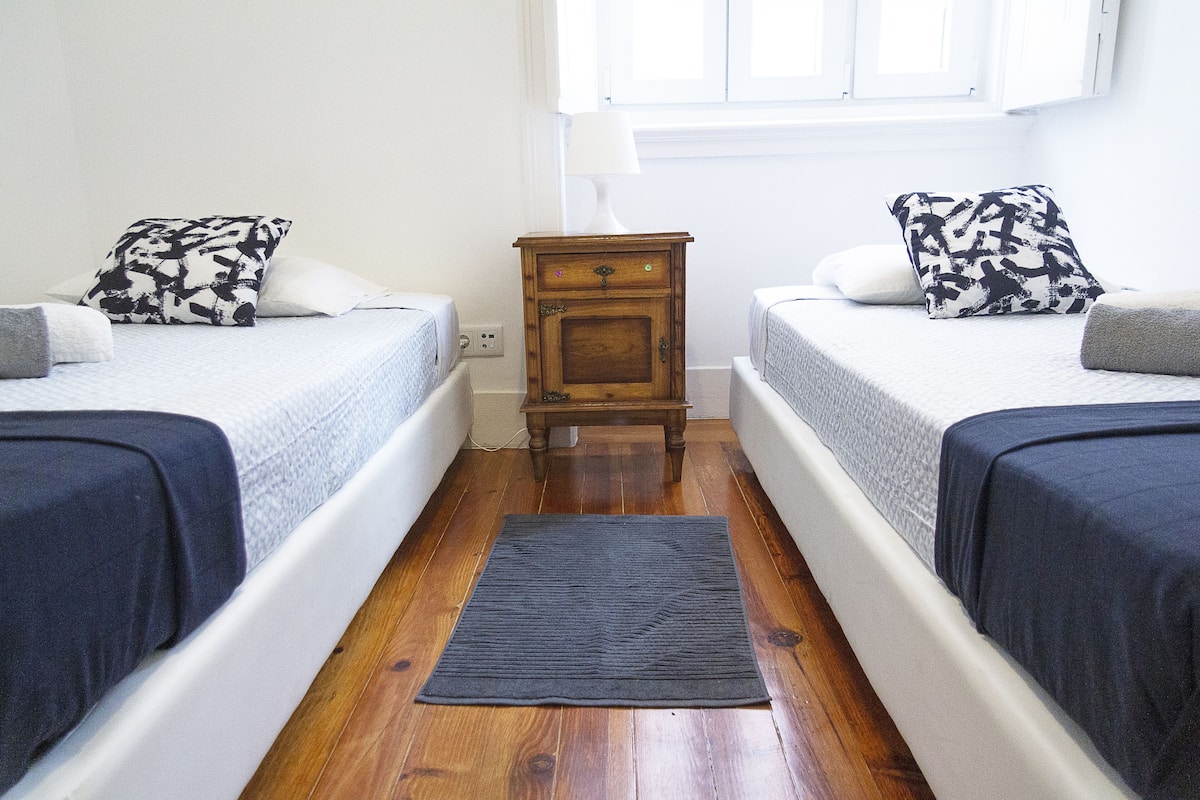 A room with two beds, a small wooden nightstand, a night lamp, and a dark rug on a wooden floor.