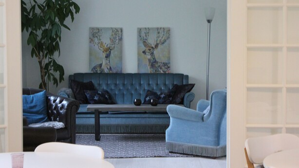 A living room with a blue sofa, a white armchair, and a coffee table. There are two paintings on the wall and a floor lamp. - Katrinelund Bed and Breakfast (Tikob)