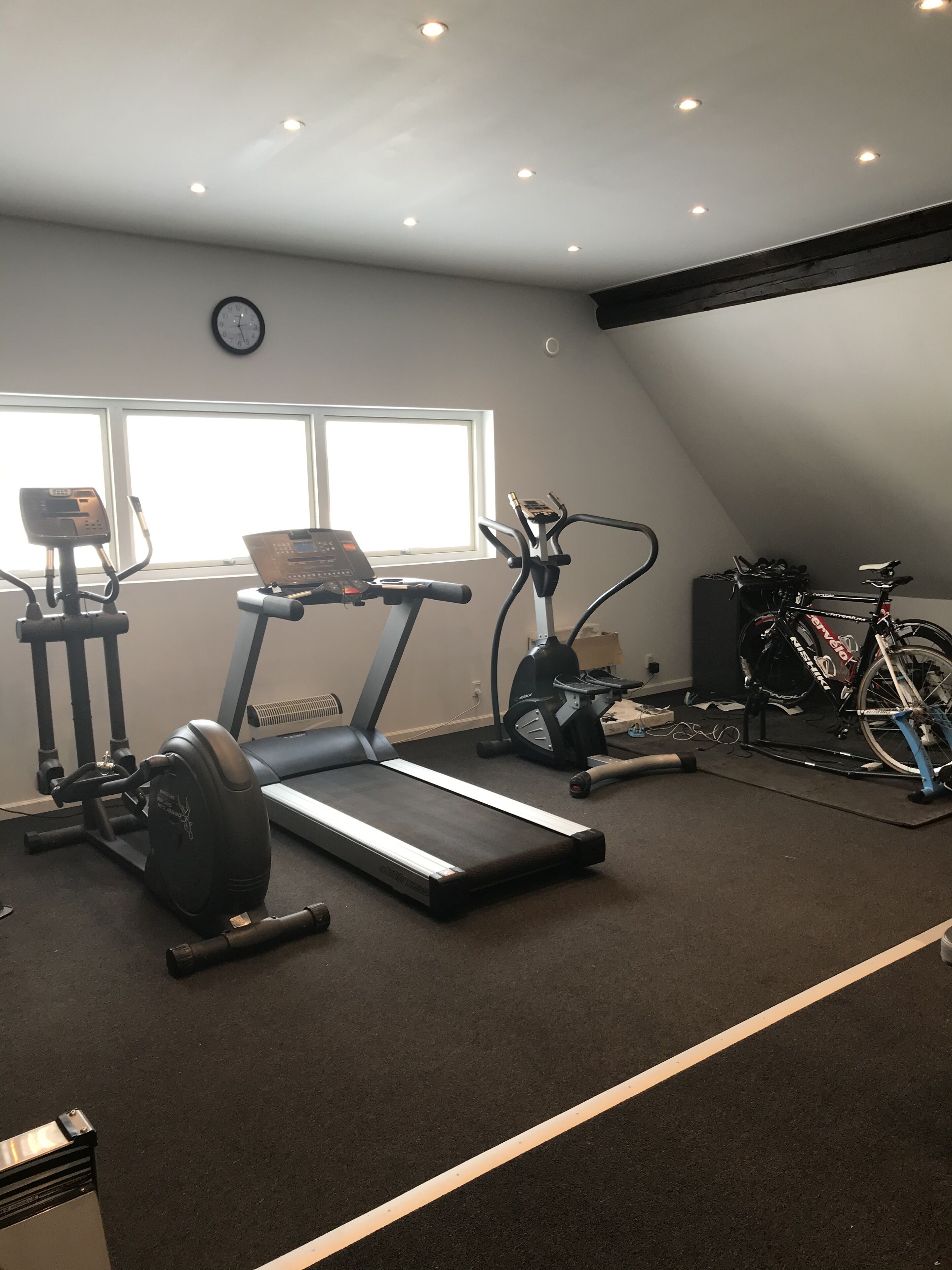 A home gym with a treadmill, elliptical, and stationary bike, a window, and a clock on the wall.