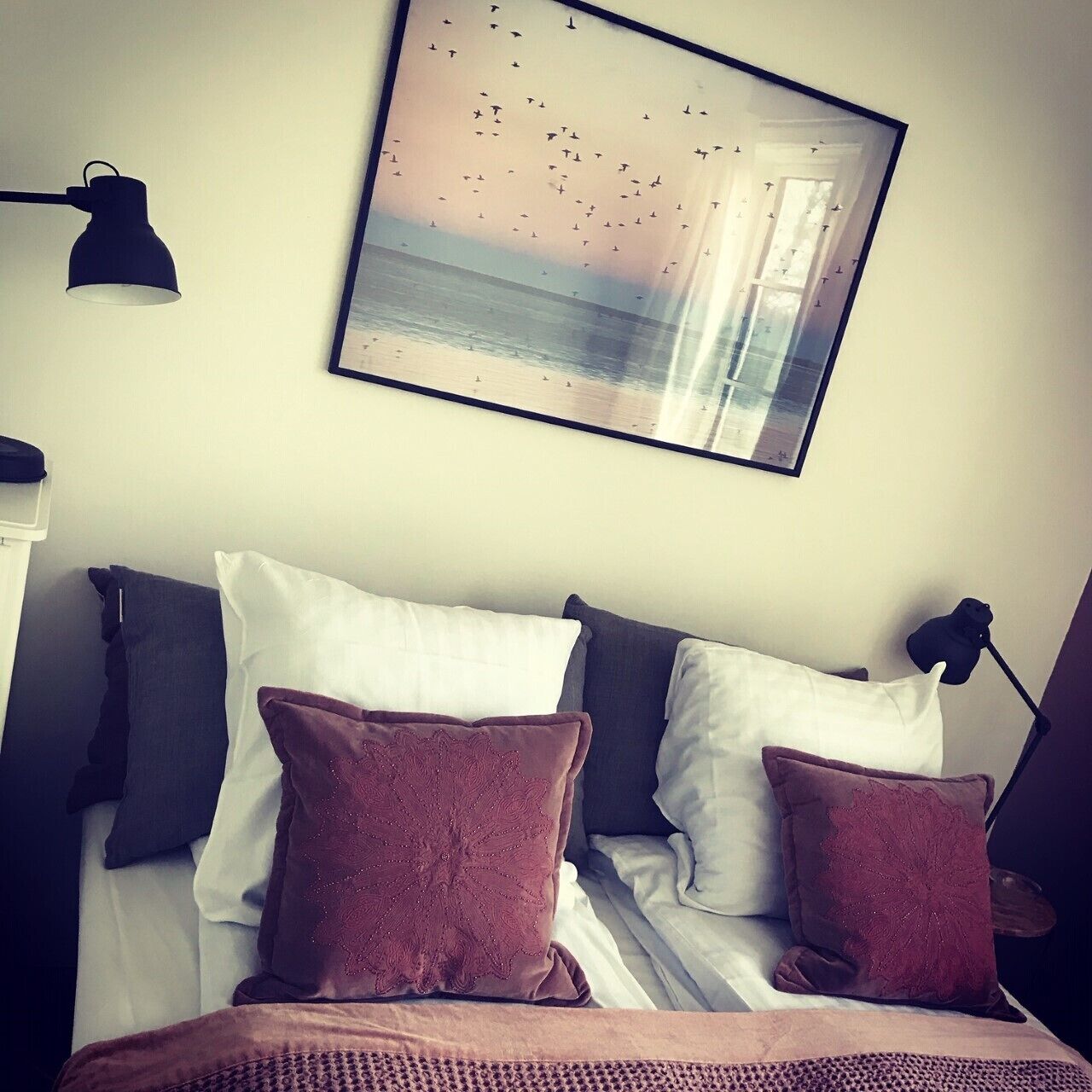 A neatly made bed with white and brown pillows, a framed picture of birds flying over water on the wall, and two black desk lamps on either side of the bed.