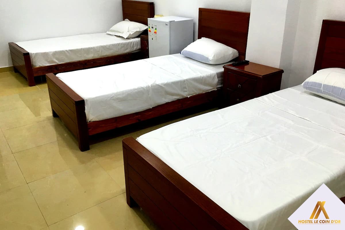 A hotel room with three single beds, each with a wooden headboard and white bedding. There is a small bedside table with a lamp on one bed.