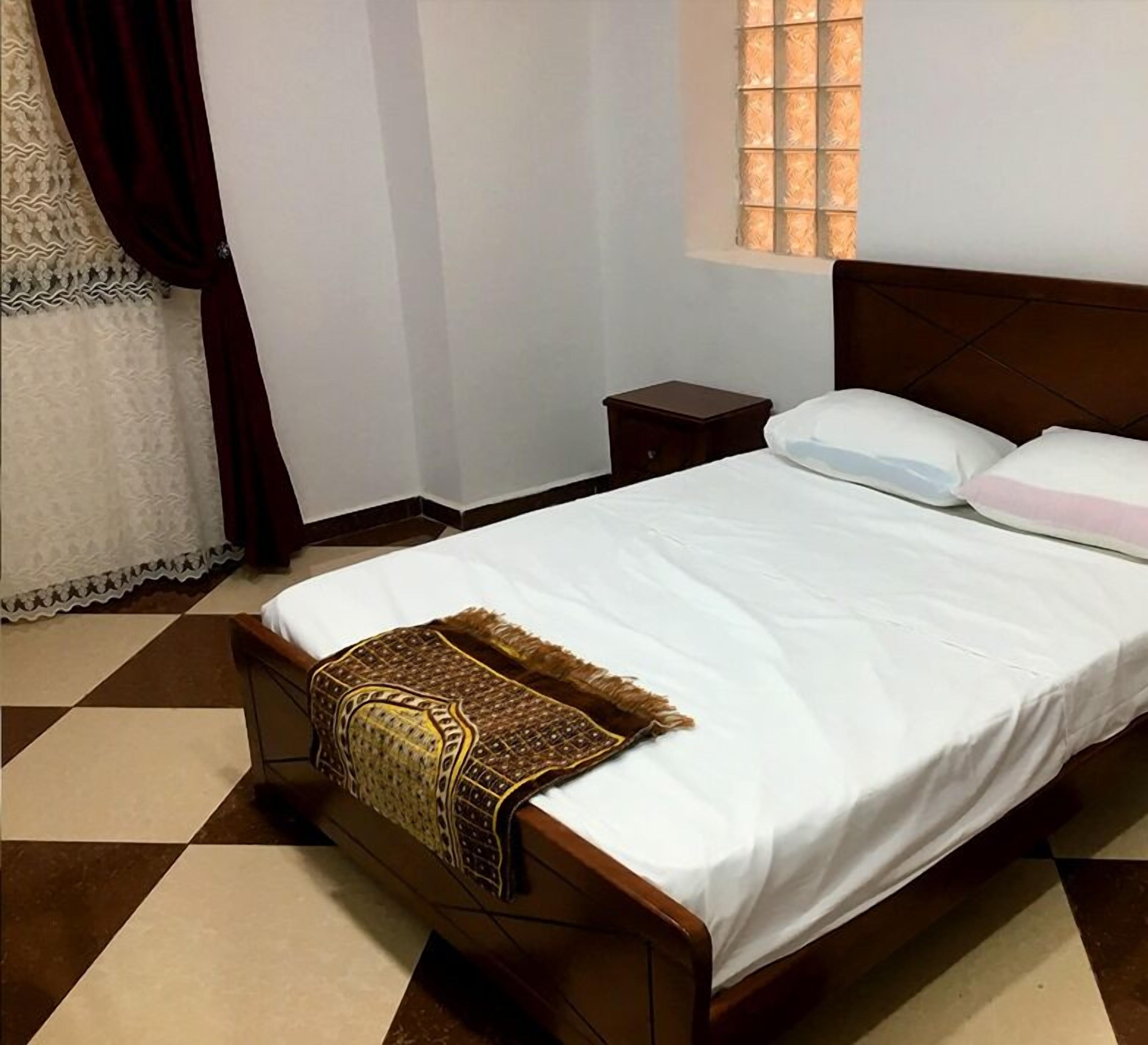 A neatly made bed with a patterned throw, a small bedside table, and a window with blinds. - Hostel Le Coin D or (Algiers)