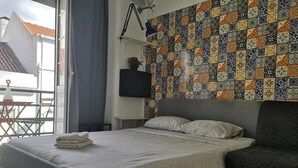 A neatly made bed with a patterned bedspread, a grey upholstered armchair, and a colorful tile wall.