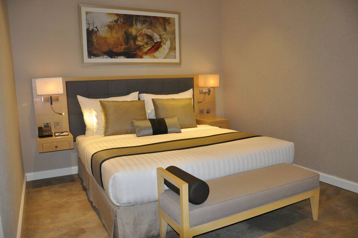 Una cama bien hecha con cabecera, dos lámparas de noche, una mesita de noche con un teléfono y un banco al pie de la cama.