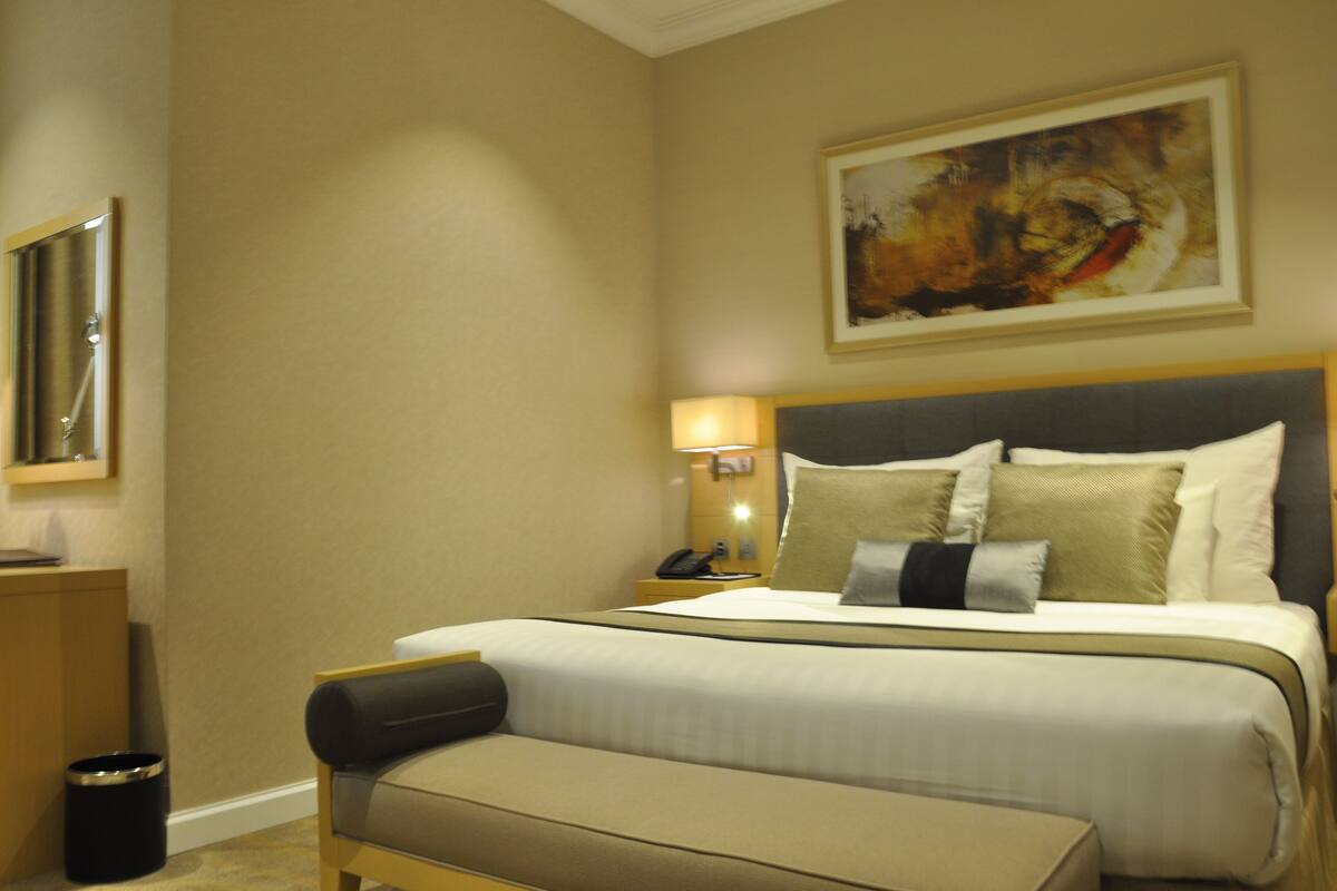 Una habitación de hotel con una cama grande, una mesita de noche con lámpara, un cubo de basura y un cuadro en la pared.