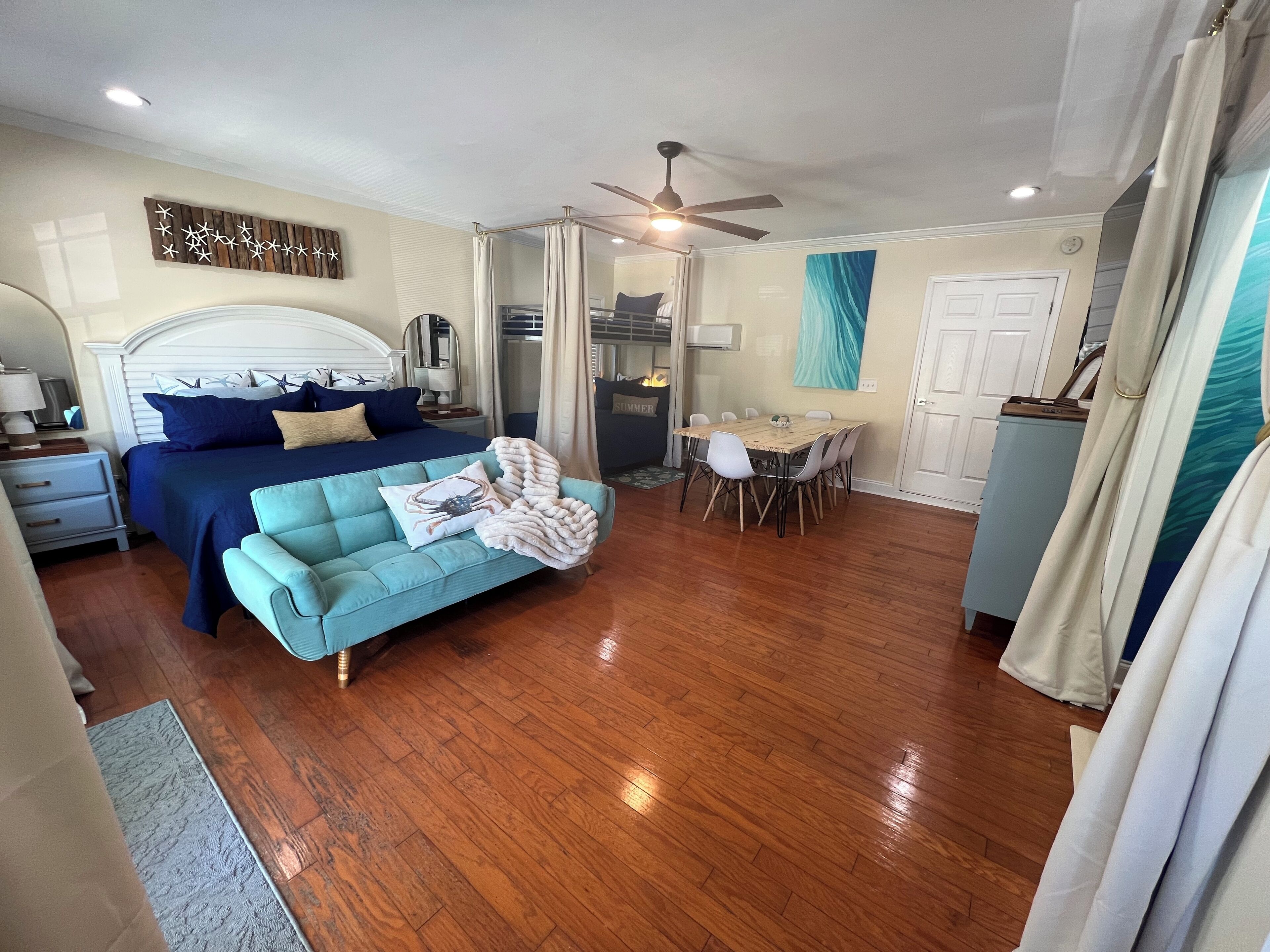 A bedroom with a bed, a sofa, a dining table with chairs, a ceiling fan, and a kitchen area.
