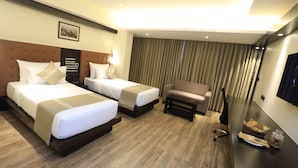 A hotel room with two beds, a sofa, a TV, and a dining table with a basket and jar of cookies. - Ocean Pearl Inn (Mangaluru)