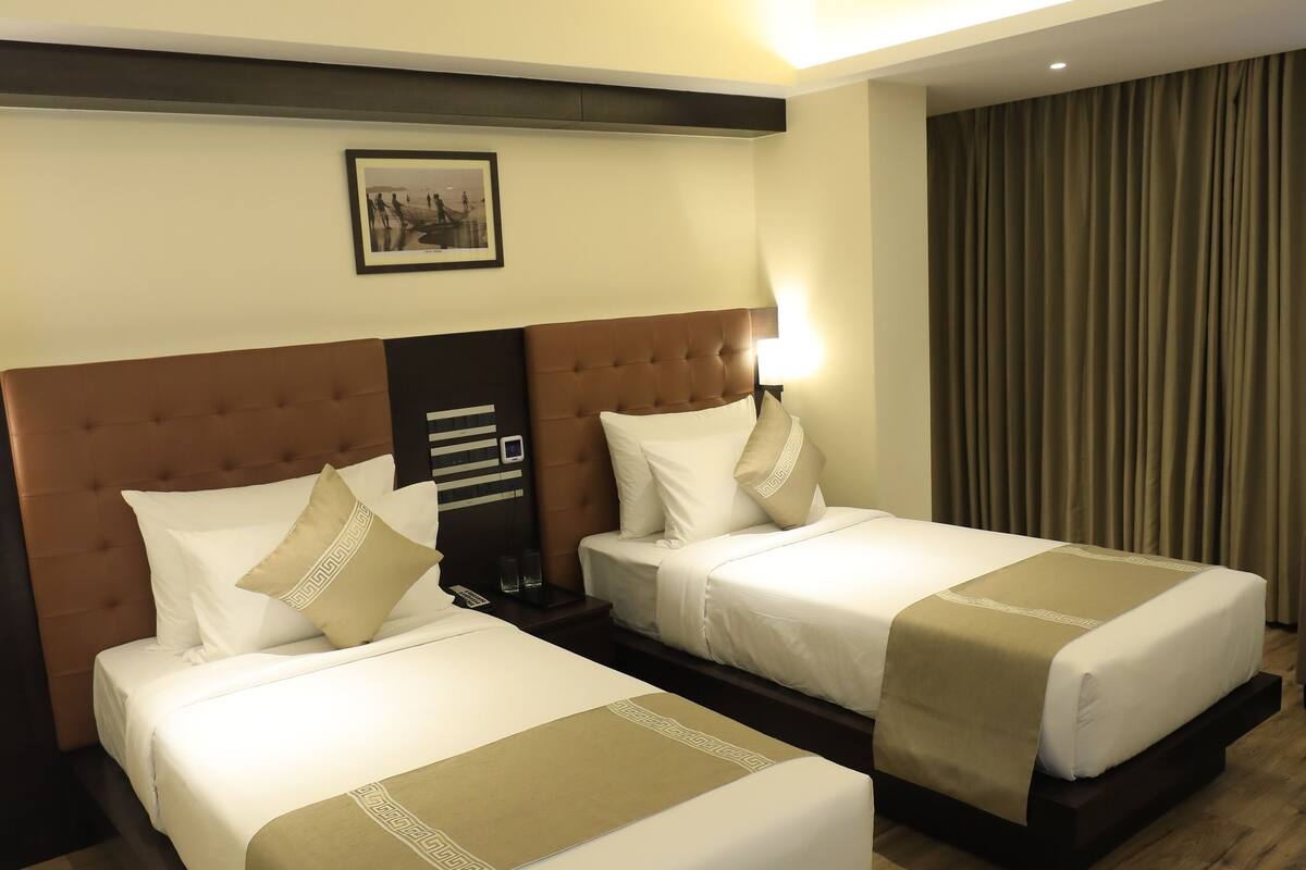 A hotel room with two beds, wooden flooring, a headboard with a light, and a framed picture on the wall.