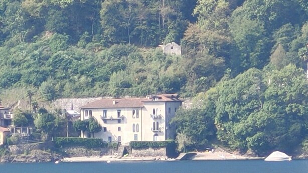 A coastal building with a balcony, surrounded by dense greenery and a body of water.