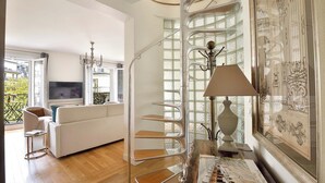 A modern living room with a white sofa, a TV, a chandelier, and a spiral staircase with a lamp. - Incredibly Luxurious Bright 2 Bed with Balcony and Wow Eiffel View (Paris)