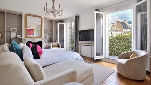 A spacious bedroom with a large bed, a television, a chandelier, and a view of the city through open windows. - Incredibly Luxurious Bright 2 Bed with Balcony and Wow Eiffel View (Paris)