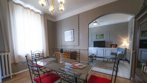 Uma área de refeições com mesa de vidro, cadeiras de metal e almofadas vermelhas. É visível uma cozinha ao fundo, com uma televisão e um quadro na parede.