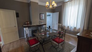 Uma sala de jantar com mesa de vidro, quatro cadeiras, um candelabro, uma cozinha ao fundo e uma porta que dá para outro espaço.