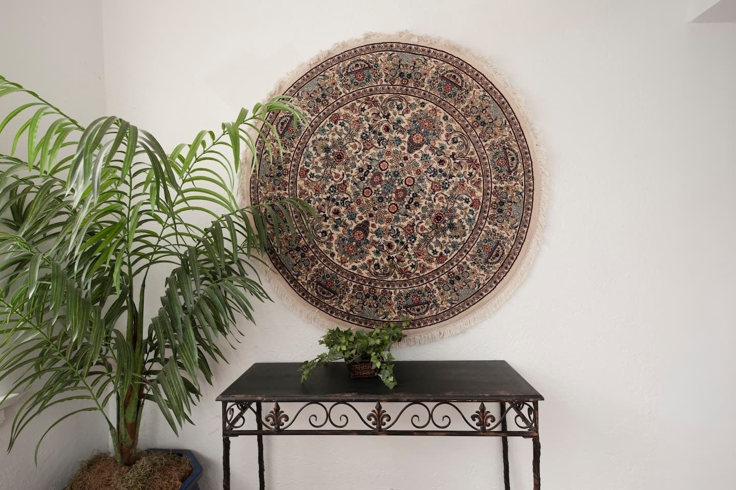 Une tapisserie ronde et richement décorée est accrochée au mur, à côté se trouve une plante en pot et une petite table sur laquelle est posée une autre plante.