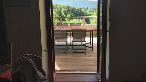 Une porte ouverte donne sur une terrasse où se trouvent une table et des chaises, et d’où l’on peut admirer un paysage boisé.