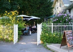 A garden entrance with a white picket fence, a signboard, and people dining under umbrellas.