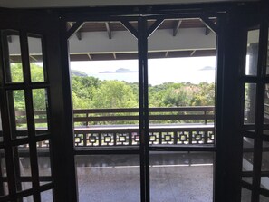 A view from a balcony with a wooden railing, overlooking a lush green landscape and a body of water with distant islands. - Tradewinds- quiet home overlooking the ocean (Bathway)