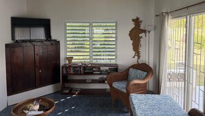 Ein Zimmer mit hoher Decke, einem Fernseher auf einer Holzkommode, einem Bücherregal, einem Korbstuhl, einem gemusterten Sofa und einem Fenster mit Jalousien.