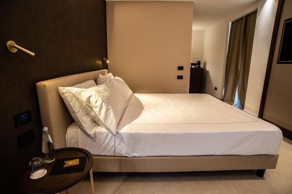 Una habitación de hotel moderna con una cama grande, una mesita de noche con una botella y vasos, y un panel de control en la pared.