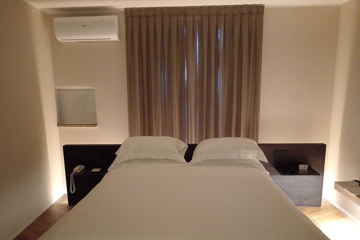 A hotel room with a large bed, a bedside telephone, a small table, and a wall-mounted air conditioning unit.